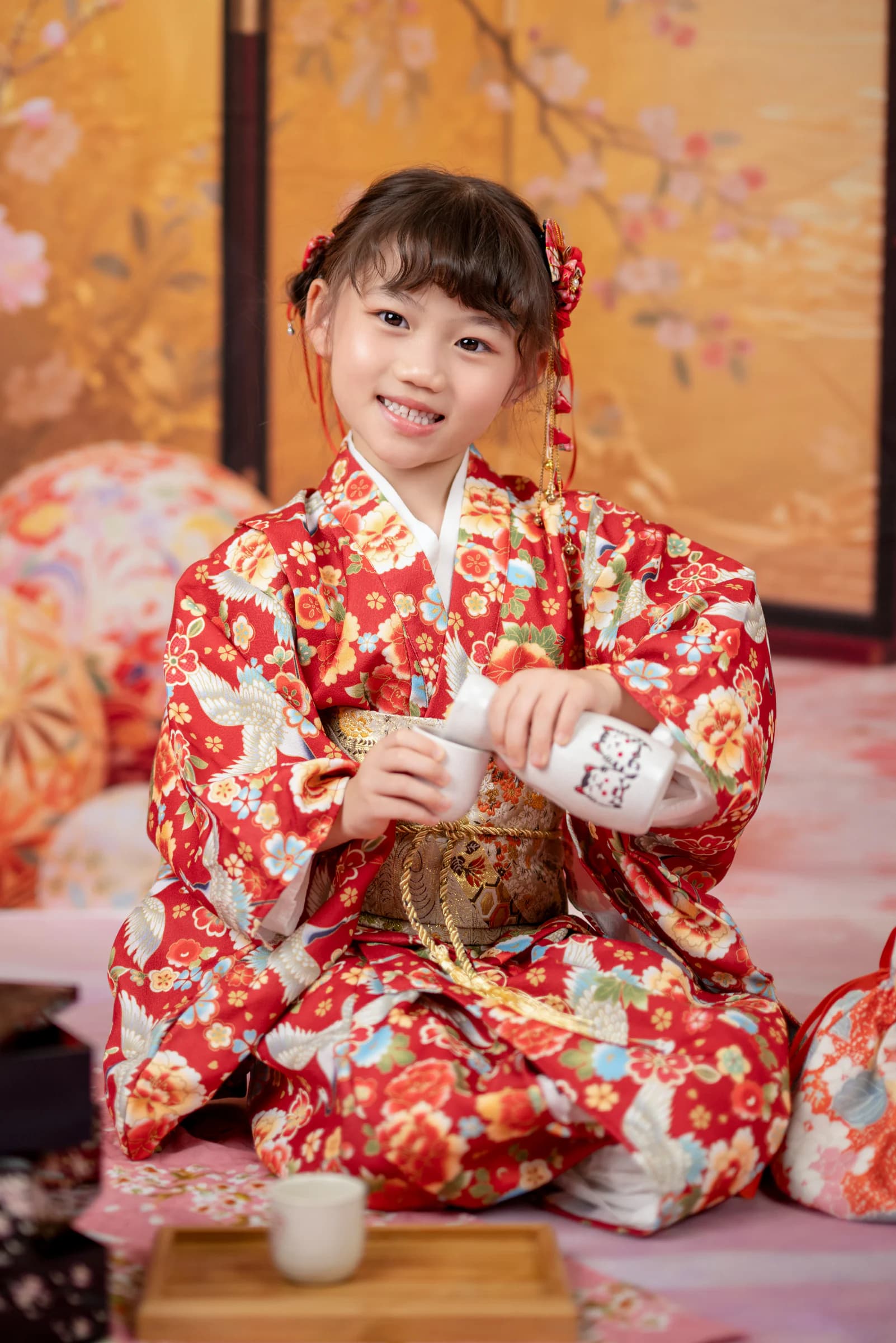 女孩穿紅色花卉和服、戴紅花髮飾,跪坐在金色櫻花屏風前笑着倒茶
